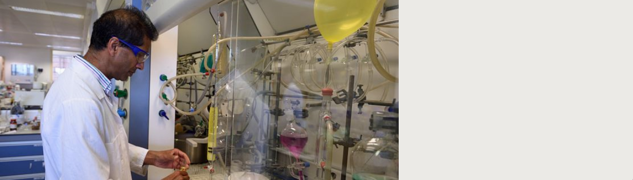 A man in goggles and a lab coat looks at some complicated laboratory apparatus containing fluids of different colours.